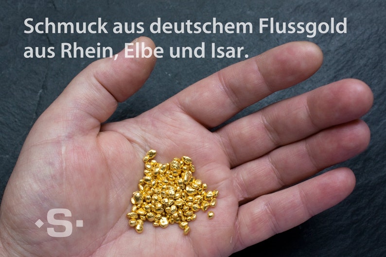 May include: A hand holding a pile of small, shiny gold nuggets. The text "Schmuck aus deutschem Flussgold aus Rhein, Elbe und Isar." is visible above the hand.