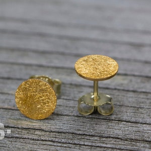 May include: A pair of gold-toned stud earrings with a textured, circular design. The earrings are on a wooden surface.