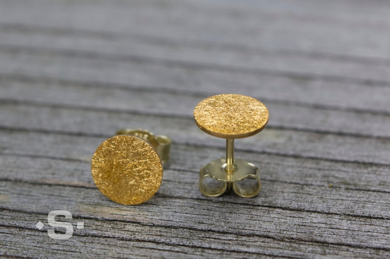 May include: A pair of gold-toned stud earrings with a textured, circular design. The earrings are on a wooden surface.