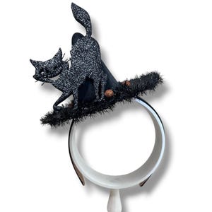 May include: A black headband featuring a miniature witch hat adorned with a glittery black cat silhouette. The hat brim is trimmed with black tinsel and small copper-colored spheres, creating a festive accessory for Halloween celebrations.