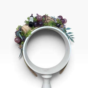 Puede incluir: Una diadema decorada con uvas moradas artificiales, follaje verde, botellas de vino en miniatura y corchos. El arreglo está fijado a una banda circular, con texturas y colores realistas que evocan un tema inspirado en los viñedos.