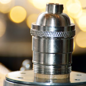 May include: A close-up of a silver-colored lamp holder. The lamp holder has a textured design around the middle and a threaded base. The background is blurred with warm, yellow bokeh lights.
