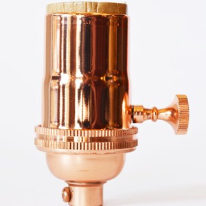 May include: A copper-colored lamp socket with a textured, ridged base and a side switch. The socket has a cylindrical body and a small, round knob. The top of the socket has a cardboard insert.