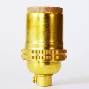 May include: A gold-colored metal light bulb socket with a brown cardboard insulator on top. The socket has a threaded base and a ribbed ring around the middle.
