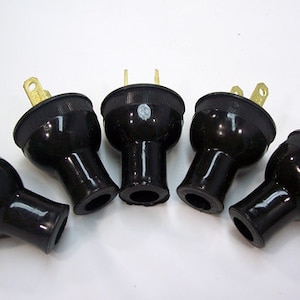 May include: Five black electrical plugs with gold-colored prongs are arranged against a white background. The plugs have a rounded, tapered shape with a ribbed texture near the top.