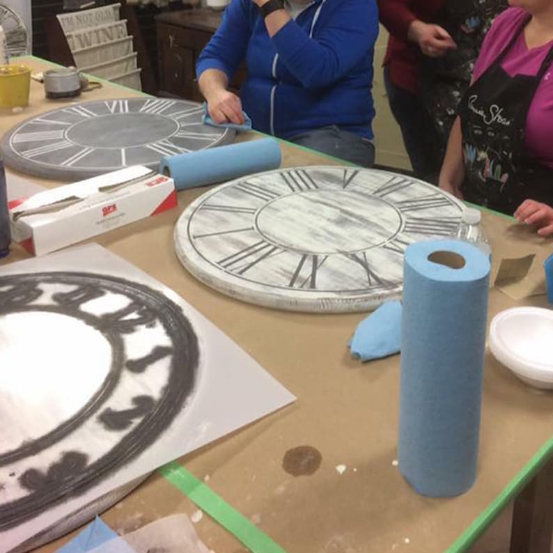 Puede incluir: Tres caras de reloj sin terminar sobre una mesa con toallas de papel azules, pinceles y un rollo de cinta de pintor. Las caras de los relojes son blancas con n&uacute;meros romanos negros.
