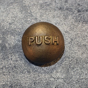 May include: A round, gold-colored door push plate with the word "PUSH" embossed on the surface.