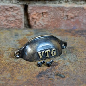 May include: A vintage-style cup pull handle with a black finish and gold lettering that reads "VTG". The handle comes with two black screws.