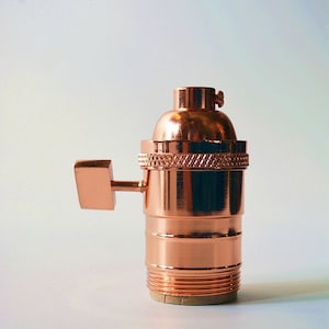 May include: A copper-colored light bulb socket with a side switch. The socket has a threaded base and a smooth, shiny finish.