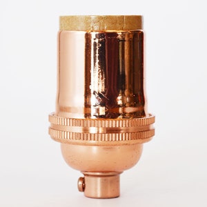 May include: A copper-colored light bulb socket with a brown cardboard insulator on top. The socket has a threaded base and a decorative ribbed design.