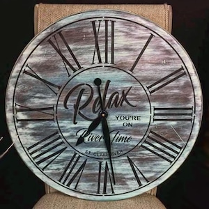 Puede incluir: Una esfera de reloj redonda de madera con n&uacute;meros romanos y una aguja de reloj negra. La esfera del reloj est&aacute; pintada en tonos azules y marrones con un aspecto desgastado. El texto "Relax You're On River Time" est&aacute; impreso en la esfera del reloj.