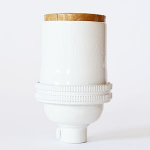 May include: A white ceramic lamp socket with a brown cork top. The socket has a cylindrical body with a ridged band near the base and a rounded bottom. The design is simple and minimalist, suitable for various lamp styles.