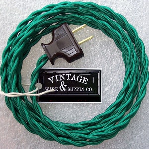 May include: A coil of emerald green twisted electrical wire with a brown plug. A black and white tag with the text "VINTAGE WIRE & SUPPLY CO." is attached to the wire with a white string. The wire is coiled in a circular shape.