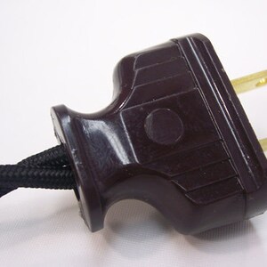 May include: Close-up of a brown electrical plug with two brass prongs. The plug has a textured, dark brown plastic housing and is connected to a black, braided electrical cord. The background is plain white.