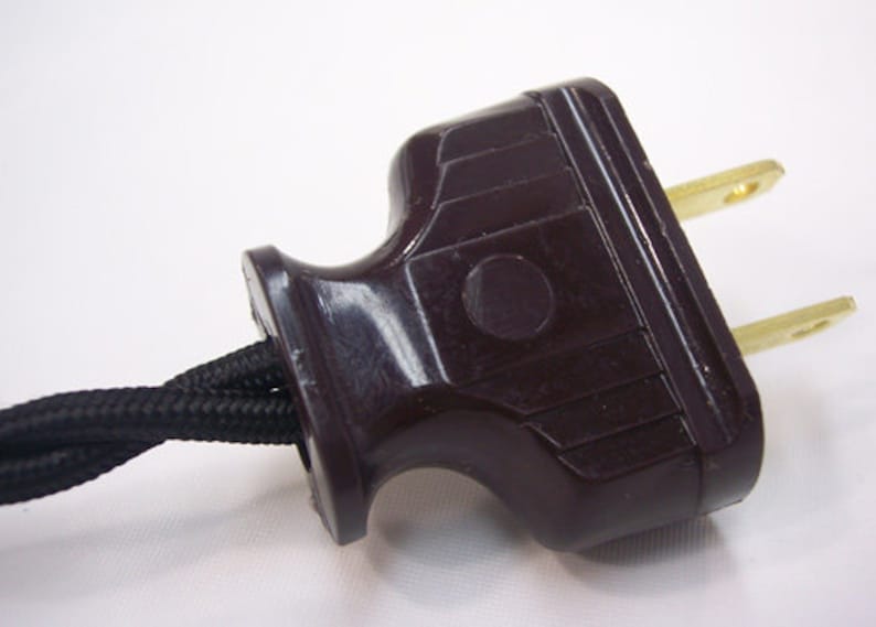 May include: Close-up of a brown electrical plug with two brass prongs. The plug has a textured, dark brown plastic housing and is connected to a black, braided electrical cord. The background is plain white.