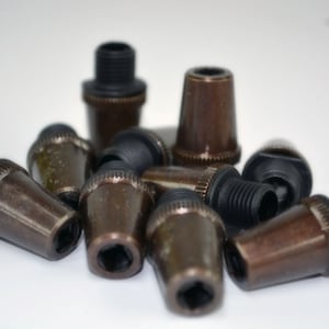 May include: A selection of bronze-coloured lamp finials with black threaded bases. The finials have a hexagonal opening and a tapered design. The objects are scattered on a white surface.