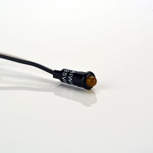 May include: A small, black, round indicator light with a yellow lens. The light has a wire attached to it and the text "AIWA 25V" printed on the side.