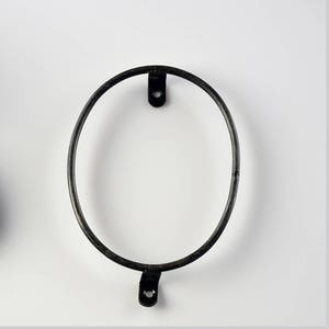 May include: Black metal oval ring with two small black metal hooks on the top and bottom. The ring is hanging on a white wall.