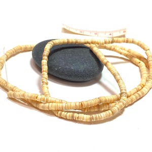 May include: A strand of small, light tan shell beads is draped over a dark gray stone. A measuring tape is in the background. The beads are strung together, creating a necklace or bracelet.