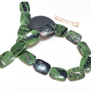 Pu&ograve; includere: Una collana di perline rettangolari, ciascuna di circa 2.5 cm di lunghezza, con sfumature verdi, nere e rosa. Le perline sono infilate, mostrando un aspetto di pietra naturale. Una pietra nera e un righello sono anche nell'immagine.
