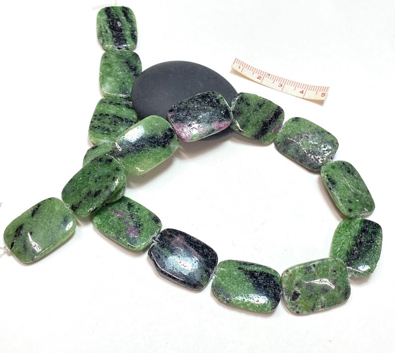 Pu&ograve; includere: Una collana di perline rettangolari, ciascuna di circa 2.5 cm di lunghezza, con sfumature verdi, nere e rosa. Le perline sono infilate, mostrando un aspetto di pietra naturale. Una pietra nera e un righello sono anche nell'immagine.