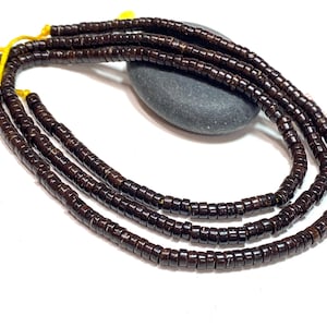 May include: Strands of dark brown, rectangular beads, likely for jewelry making, are displayed against a white background. The beads are strung on yellow thread. A gray stone rests behind the strands.
