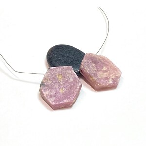 May include: Two rough-cut, hexagonal pink gemstones and a dark gray stone are strung on a thin, silver-colored wire. The gemstones have a textured surface and varying shades of pink. The background is white.
