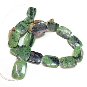 Pu&ograve; includere: Un filo di perline rettangolari di zoisite. Le perline sono di un verde vibrante con inclusioni nere e rosa. Le perline sono infilate su un cordino sottile e trasparente e sono esposte su un pezzo di legno.