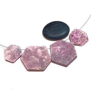 May include: A necklace featuring five hexagonal, light purple gemstones and a dark gray oval stone. The gemstones have a rough, textured surface and are strung on a thin, silver-colored wire. The background is plain white.