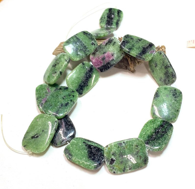 Pu&ograve; includere: Una collana di perline rettangolari di zoisite. Le perline sono un mix di verde, nero e rosa. Le perline sono infilate su un cordino sottile e trasparente. Le perline sono lisce e lucide.
