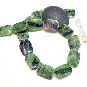 Pu&ograve; includere: Una collana di perline rettangolari verdi, nere e rosa. Le perline sono infilate insieme, con una pietra grigio scuro e un righello sullo sfondo. Le perline sono lunghe circa 2,5 cm.