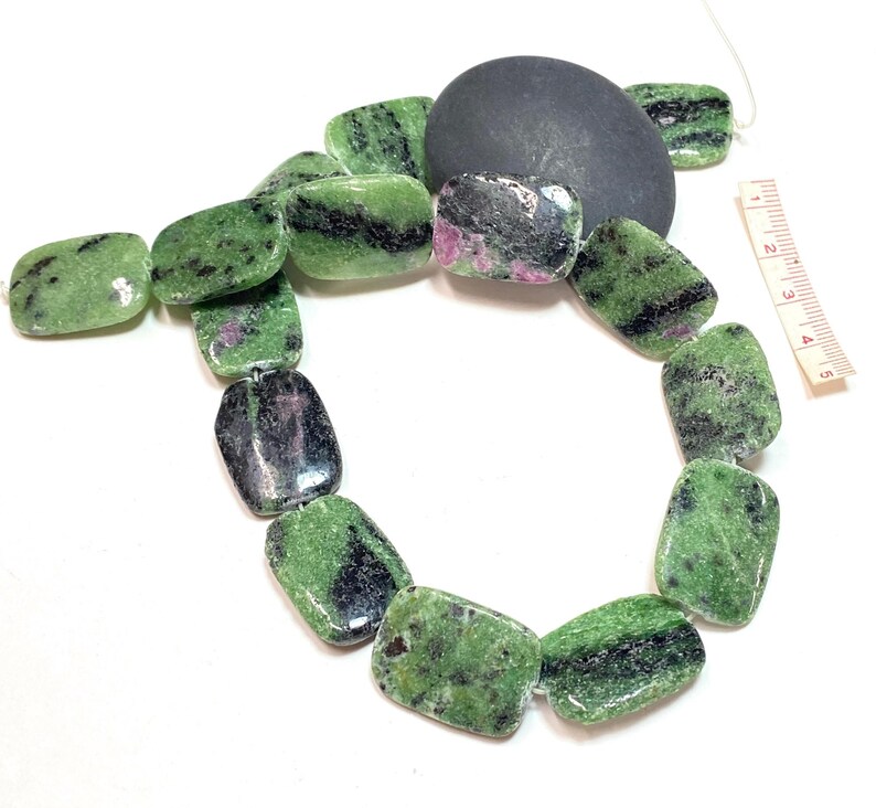 Pu&ograve; includere: Una collana di perline rettangolari verdi, nere e rosa. Le perline sono infilate insieme, con una pietra grigio scuro e un righello sullo sfondo. Le perline sono lunghe circa 2,5 cm.