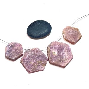 May include: A collection of raw, hexagonal-shaped pink gemstones strung on a wire, alongside a smooth, dark blue oval stone. The gemstones have a rough, textured surface and varying shades of pink.