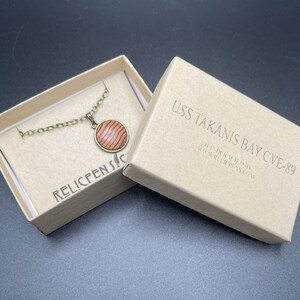 May include: A wooden pendant necklace with a brown and red wood grain design. The pendant is in a small box with the text "USS TAKANIS BAY CVE-89 HANDCRAFTED BY WWW.RELICPENS.COM" printed on the box.
