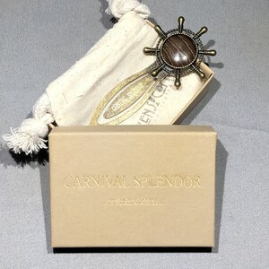 May include: A gold-toned bookmark featuring a wooden ship's wheel design. The bookmark is presented in a small, brown box with the text "CARNIVAL SPLENDOR" printed on the top.