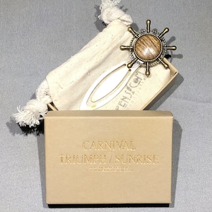May include: A white metal bookmark with a wooden ship's wheel charm. The bookmark is in a small brown box with the text "Carnival Triumph / Sunrise Handmade Jewelry www.thegoldenrose.com".