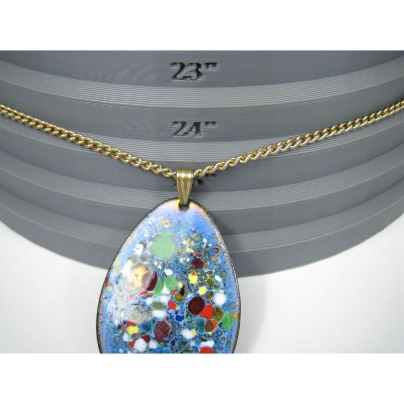 Puede incluir: Un collar de cadena dorada con un colgante azul en forma de l&aacute;grima. El colgante presenta un dise&ntilde;o de esmalte colorido con detalles en rojo, verde y amarillo. El collar se muestra sobre una superficie gris.