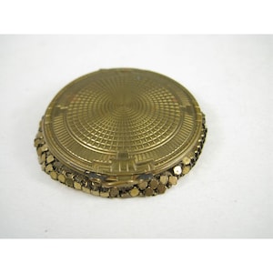 May include: A round, brass-toned compact mirror featuring a textured, geometric grid pattern on the lid. The sides of the compact are encased in a flexible, metallic mesh material composed of small, interlocking square-shaped links.