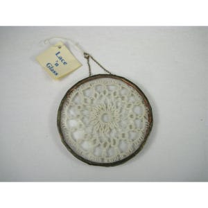 Pu&ograve; includere: Un centrino rotondo in pizzo color crema, incorniciato in un anello di metallo argentato. Il pizzo presenta un motivo simmetrico e dettagliato. Una piccola etichetta attaccata in alto recita "Lace 'n Glass". Il centrino &egrave; appeso a una delicata catena.