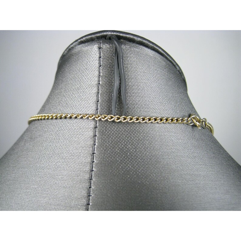 Puede incluir: Un collar de cadena de color dorado expuesto sobre un maniqu&iacute; gris. La cadena es de estilo eslab&oacute;n de bordillo, con un cierre sencillo. El collar se muestra desde la parte posterior, destacando el dise&ntilde;o de la cadena y los detalles del cierre.
