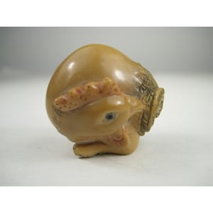 May include: A small, detailed sculpture of a bird, possibly a chicken or rooster, carved from a light brown material. The bird is in a curled position, with its head turned and beak tucked in. The sculpture has a textured appearance.