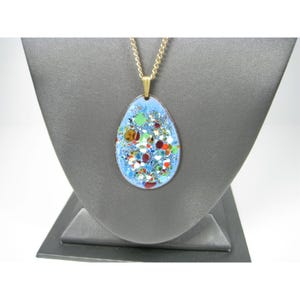 Puede incluir: Un collar dorado con un colgante ovalado. El colgante tiene un fondo de esmalte azul claro y un dise&ntilde;o moteado colorido. El collar se exhibe en un expositor de joyer&iacute;a gris.