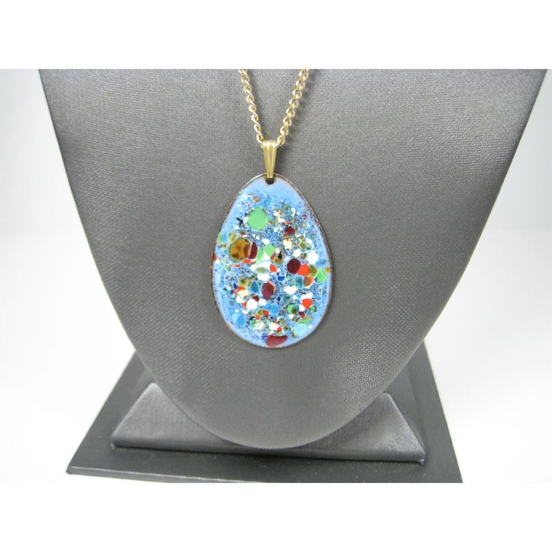 Puede incluir: Un collar dorado con un colgante ovalado. El colgante tiene un fondo de esmalte azul claro y un dise&ntilde;o moteado colorido. El collar se exhibe en un expositor de joyer&iacute;a gris.