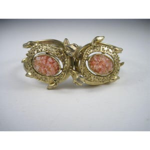 May include: A gold-toned metal cuff bracelet featuring two turtle-shaped motifs. Each turtle shell is textured with a granular surface and contains an oval-shaped, light pink, crystalline stone inlay at the center of the shell design.