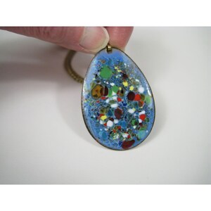 Puede incluir: Un colgante en forma de l&aacute;grima con una superficie de esmalte azul, que presenta un dise&ntilde;o moteado colorido de rojo, verde, amarillo y blanco. El colgante tiene un borde y una cadena de metal dorado, creando una pieza de joyer&iacute;a vintage.
