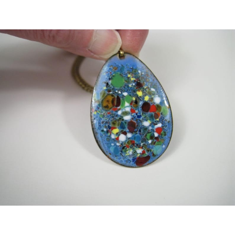 Puede incluir: Un colgante en forma de l&aacute;grima con una superficie de esmalte azul, que presenta un dise&ntilde;o moteado colorido de rojo, verde, amarillo y blanco. El colgante tiene un borde y una cadena de metal dorado, creando una pieza de joyer&iacute;a vintage.