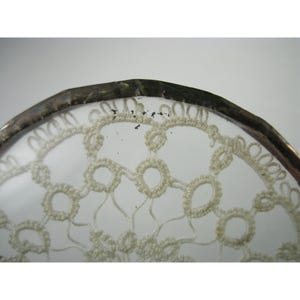 Pu&ograve; includere: Primo piano di un oggetto in vetro trasparente con un bordo in metallo color argento. Il vetro presenta un delicato motivo a pizzo color crema. Il disegno del pizzo &egrave; intricato, con forme circolari e curve ripetute.