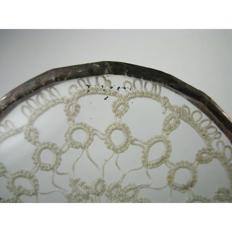 Pu&ograve; includere: Primo piano di un oggetto in vetro trasparente con un bordo in metallo color argento. Il vetro presenta un delicato motivo a pizzo color crema. Il disegno del pizzo &egrave; intricato, con forme circolari e curve ripetute.