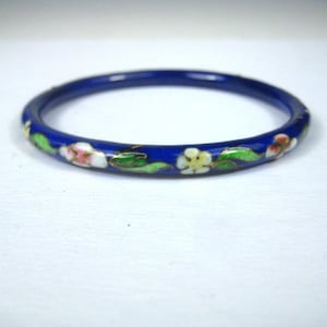 May include: A thin, circular bangle bracelet featuring a deep royal blue enamel base. The surface is decorated with a repeating pattern of small, multi-colored flowers in shades of white, pink, and yellow, accented by vibrant green leaves.