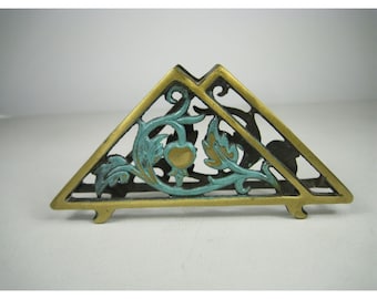 Puede incluir: Un servilletero triangular hecho de latón con un acabado dorado metálico. El marco presenta un diseño calado de hojas en espiral y un motivo de fruta central, acentuado con un acabado de pátina turquesa en los elementos florales decorativos.